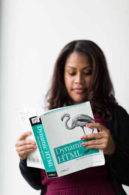 A woman with dark, shoulder-length hair and medium skin tone is standing indoors against a plain white wall, holding a book titled 'Dynamic HTML' by O'Reilly with a cover featuring a stylized illustra